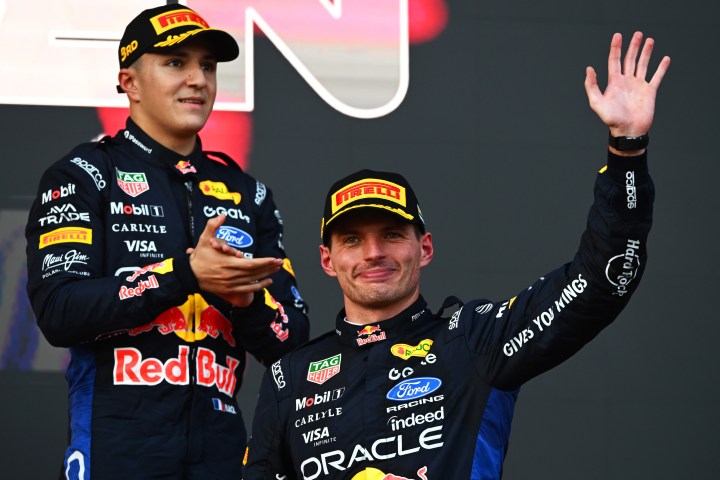 Zwei Rennfahrer auf dem Podium, einer winkt und lächelt, der andere applaudiert. Beide tragen Rennanzüge mit Sponsorenlogos.